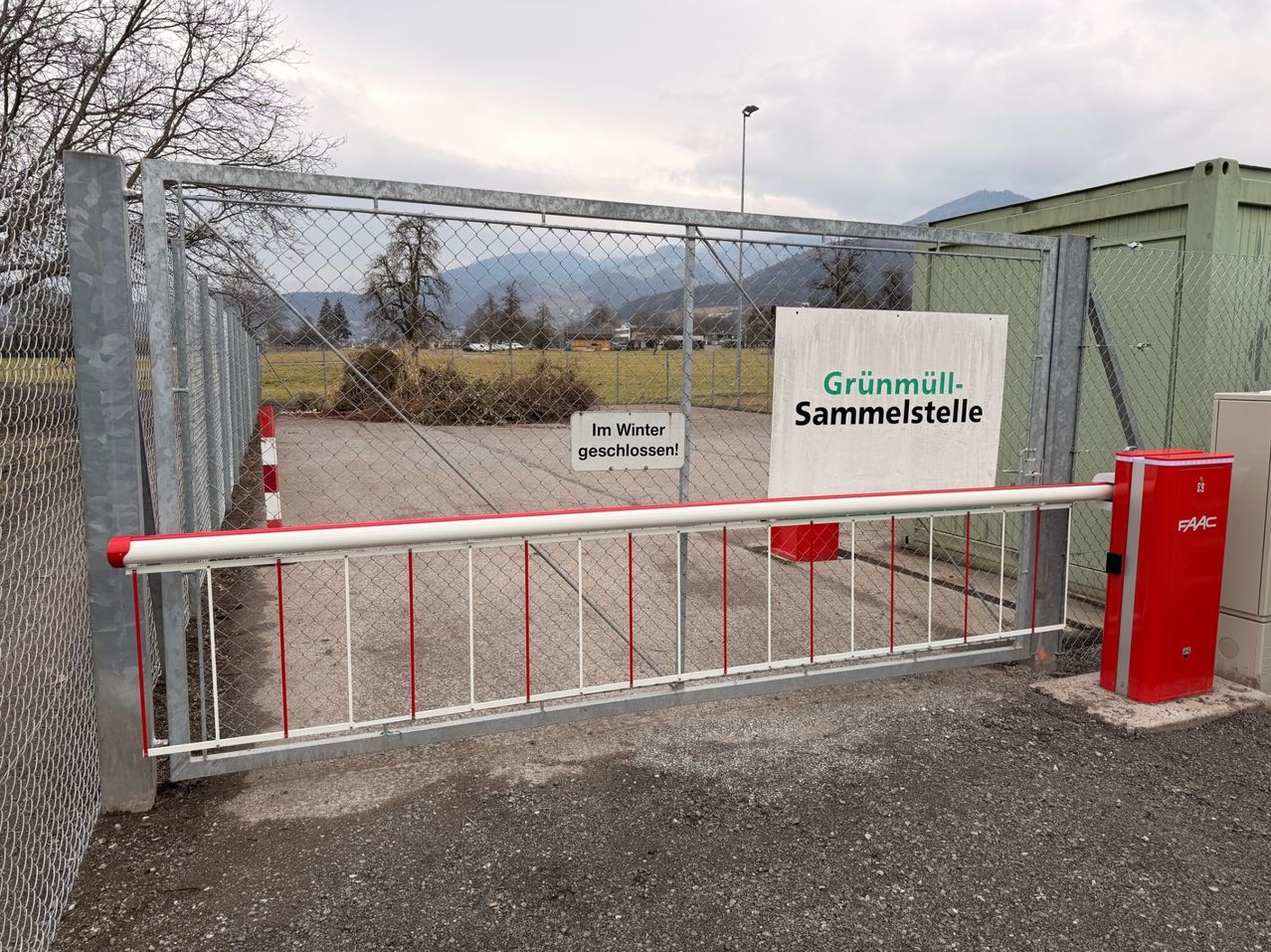 Ein geschlossenes Tor mit einem Schild, das Grünmüll-Sammelstelle und Im Winter geschlossen liest. Das Tor ist aus Metall mit roten und weißen Streifen. Dahinter befindet sich eine Grasfläche mit Bergen in der Ferne.