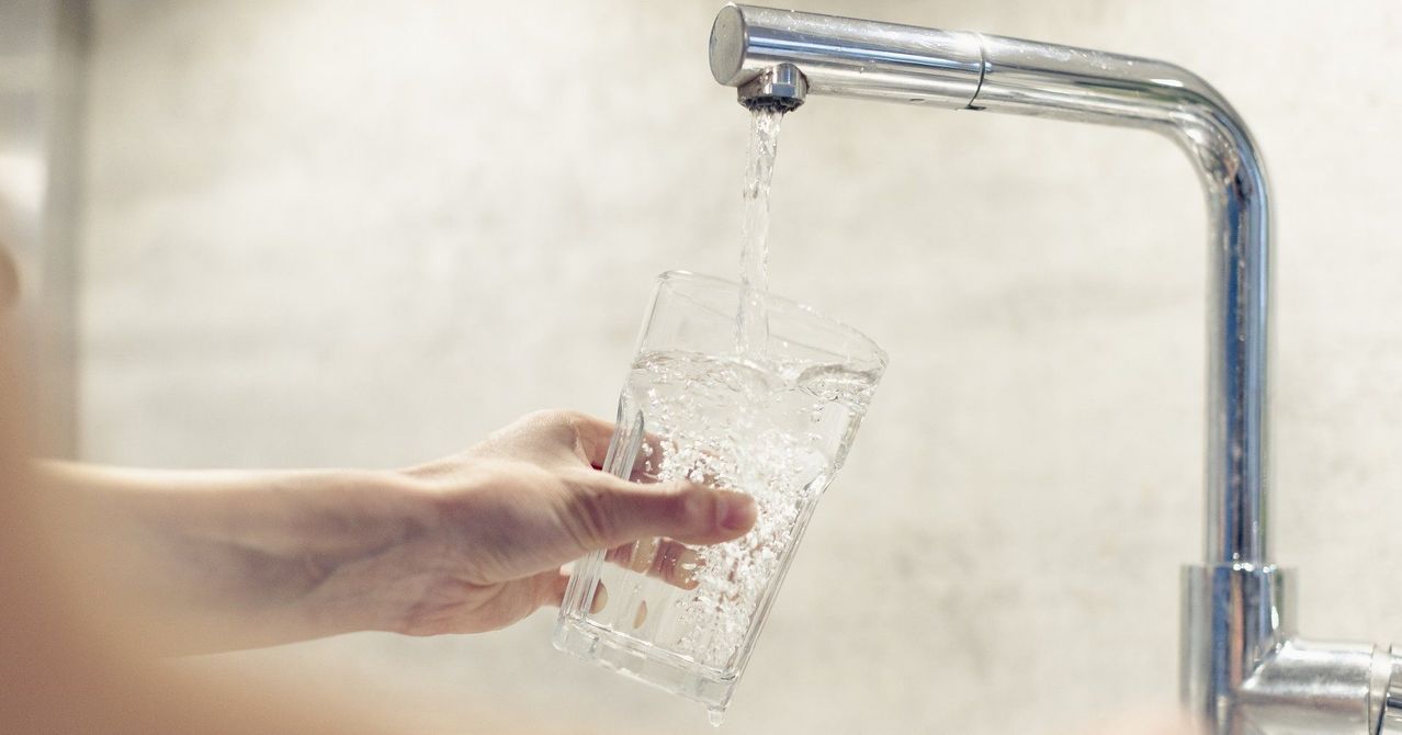 Eine Hand füllt ein klares Glas mit Wasser aus einem Edelstahlhahn. Das Wasser ist klar und es sind Blasen sichtbar, während es das Glas füllt.