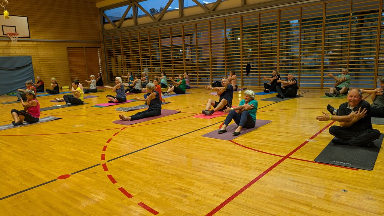 Eine Gruppe von Menschen praktiziert Yoga auf Yogamatten in einer Turnhalle. Sie sitzen in verschiedenen Positionen auf dem Boden.