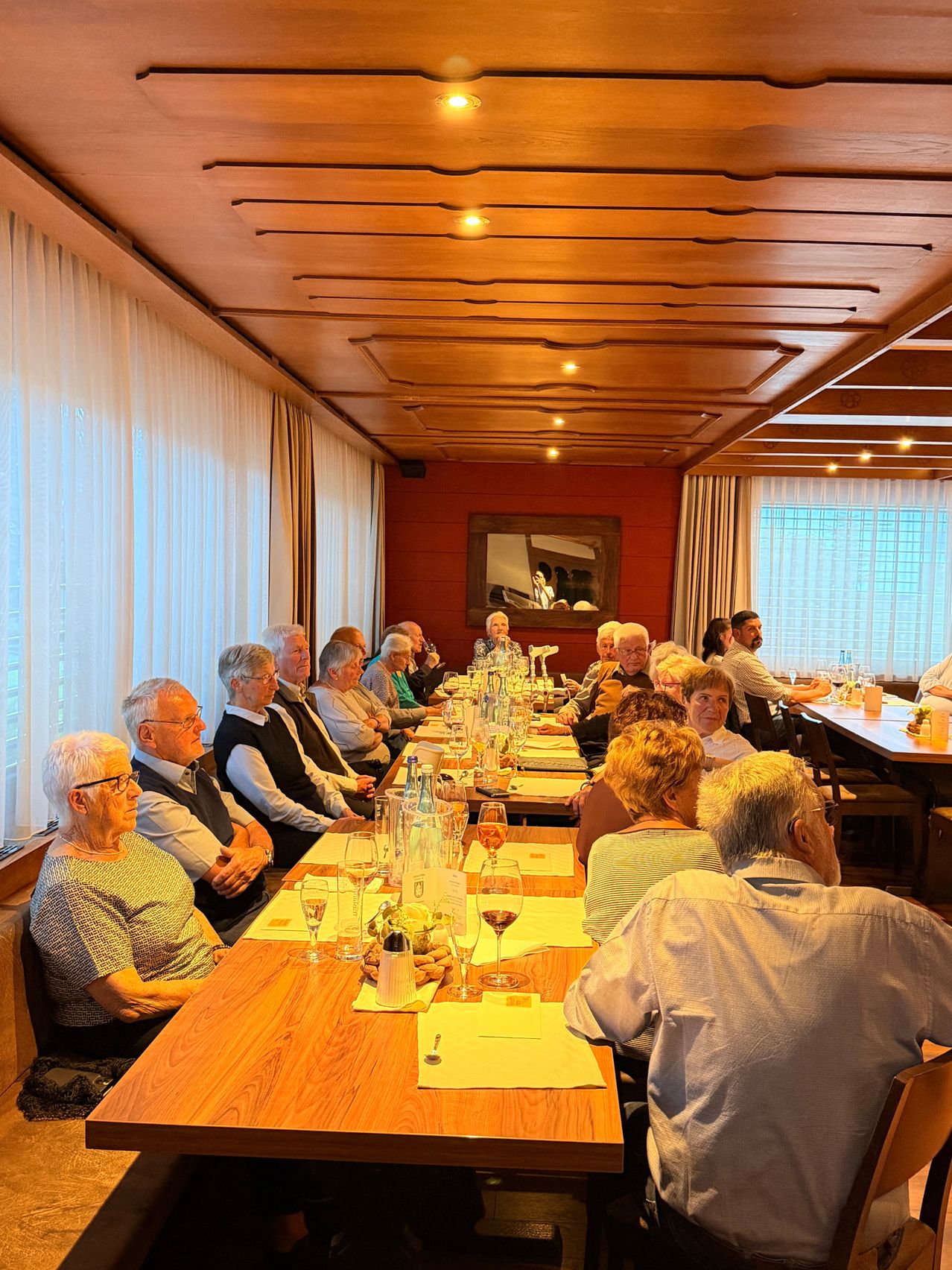 Eine Gruppe älterer Menschen sitzt um einen langen Tisch in einem Restaurant. Auf dem Tisch stehen Weingläser, Wasserflaschen und Servietten. Der Raum hat Holzdecken, rote Wände und weiße Vorhänge. An der Wand hängt ein Spiegel und ein Fenster mit Jalousien.