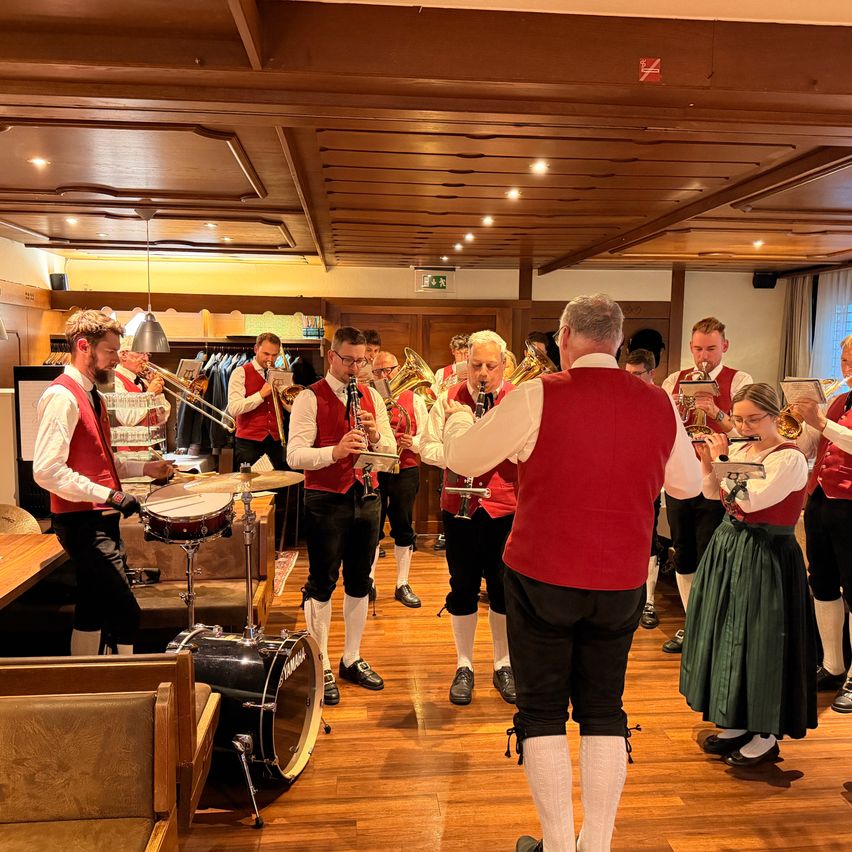Eine Gruppe von Musikern in traditioneller Kleidung spielt Instrumente auf einem Holzboden. Der Mann in der Mitte spielt Trompete.