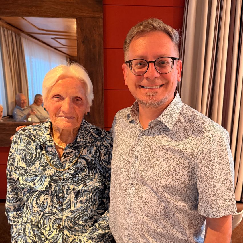 Eine ältere Frau mit weißen Haaren und einer Halskette posiert für ein Foto mit einem Mann in einer gemusterten Bluse und einer Brille vor einer roten Wand.