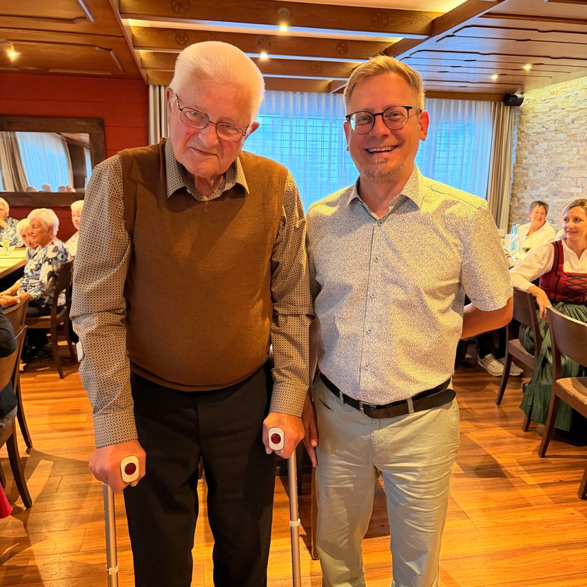 Zwei Männer stehen in einem Esszimmer, einer benutzt einen Gehstock. Der ältere Mann trägt eine Brille und eine braune Weste und lächelt. Der jüngere Mann trägt ein weißes Hemd und Khakihosen. Hinter ihnen sitzen andere Menschen an Tischen.