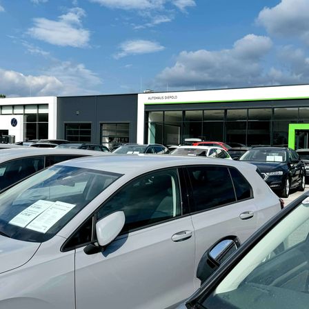 Ein Autohaus mit blauem Himmel und Wolken im Hintergrund. Viele Autos sind draußen geparkt, darunter ein silberner Wagen mit einem Schild auf der Windschutzscheibe.
