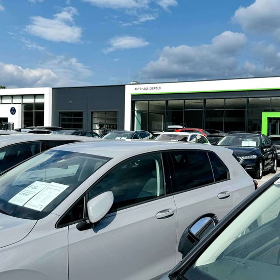 Ein Autohaus mit blauem Himmel und Wolken im Hintergrund. Viele Autos sind draußen geparkt, darunter ein silberner Wagen mit einem Schild auf der Windschutzscheibe.