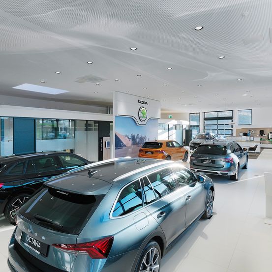 Ein Skoda-Autohaus zeigt mehrere Fahrzeuge, darunter einen silbernen Skoda Octavia, ein orangefarbenes Skoda-Auto und einen grauen Skoda Scout. Die Ausstellungsfläche hat weiße geflieste Böden und ein modernes Deckendesign.