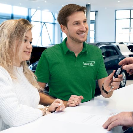 Ein Paar sitzt an einem Schreibtisch in einem Autohaus. Der Mann hält einen Autoschlüssel und scheint mit der Frau zu sprechen. Hinter ihnen sind Autos geparkt.