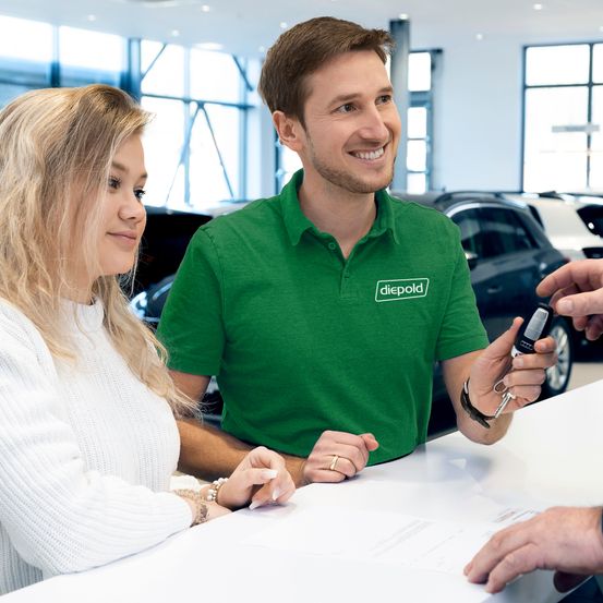 Ein Paar sitzt an einem Schreibtisch in einem Autohaus. Der Mann hält einen Autoschlüssel und scheint mit der Frau zu sprechen. Hinter ihnen sind Autos geparkt.