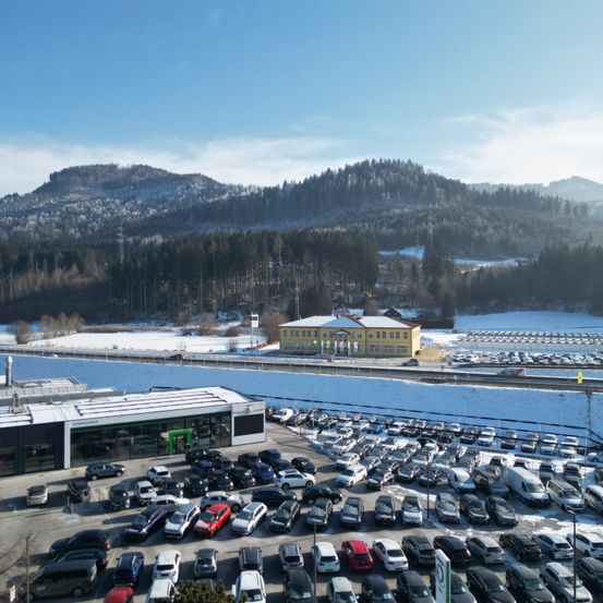 Luftaufnahme eines geschäftigen Autohauses mit vielen geparkten Fahrzeugen, einem gelben Gebäude und verschneiten Bergen im Hintergrund.