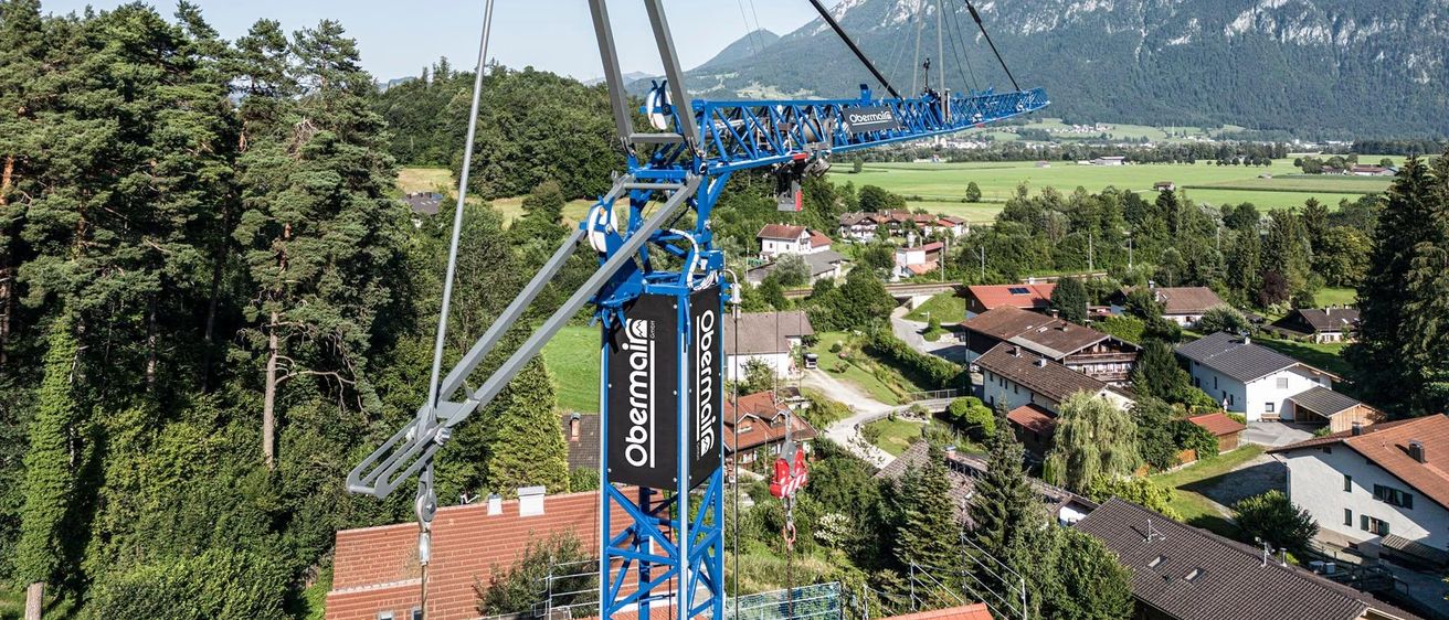 Ein blauer Baukran steht hoch über einer Stadt mit Häusern, umgeben von Bäumen und Bergen.