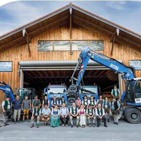 Eine Gruppe von Menschen in traditioneller Kleidung posiert vor einem Holzgebäude mit schwerem Gerät im Inneren.