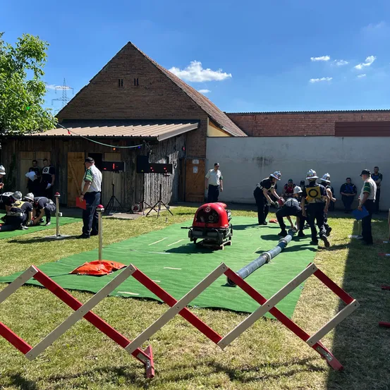 Eine Gruppe Feuerwehrleute trainiert auf einem grünen Matten mit Ausrüstung, Barrieren und einem Gebäude im Hintergrund unter einem klaren Himmel.