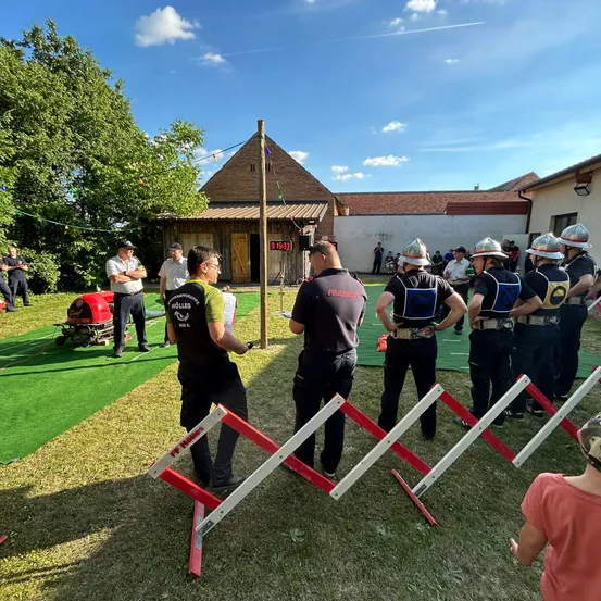 Eine Gruppe Feuerwehrleute in voller Ausrüstung steht in einer Reihe auf einem grünen Feld. Eine digitale Uhr und ein Gebäude sind hinter ihnen. In der Ferne steht eine Person und beobachtet das Ereignis.