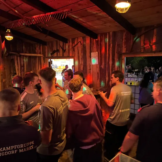 Eine Gruppe von Männern und Frauen versammelt sich in einer Bar mit Holzwänden und roten Lichtern. Einige tragen T-Shirts mit Text und Zahlen, während andere am Tresen stehen.