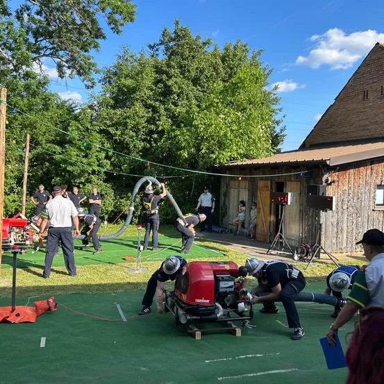 Eine Gruppe von Menschen übt draußen Brandschutzübungen. Einige halten Schläuche und Rohre, andere arbeiten an einer roten Maschine. Das Gebiet ist von Bäumen umgeben, und im Hintergrund steht ein Gebäude.