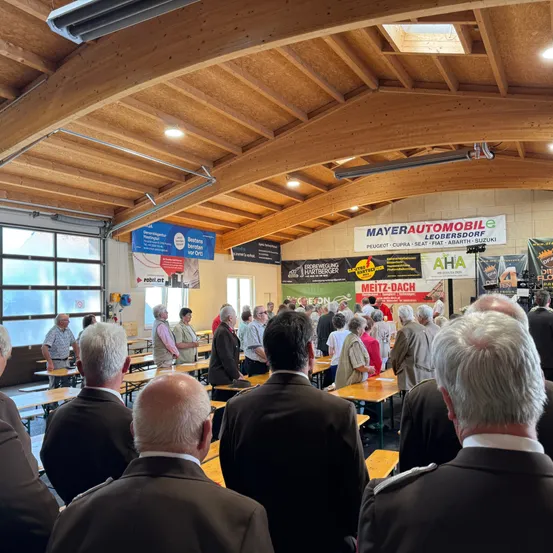 Eine Gruppe von Menschen in formeller Kleidung versammelt sich in einem Raum, wahrscheinlich für eine Besprechung. Der Raum hat Holztische und -stühle, und Banner hängen an der Wand. Es sind Lampen an der Decke montiert.