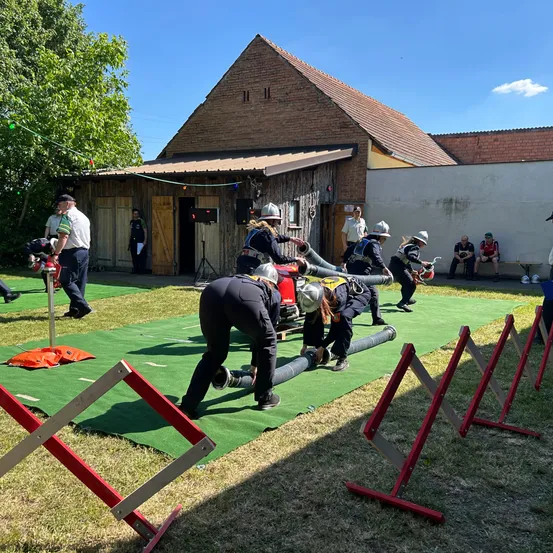 Eine Gruppe von Menschen in Feuerwehrausrüstung führt eine Trainingsübung auf einem grünen Feld durch. Barrieren und ein roter Schlauch sind auf dem Feld. Es gibt Zuschauer, die auf einer Bank im Hintergrund sitzen.