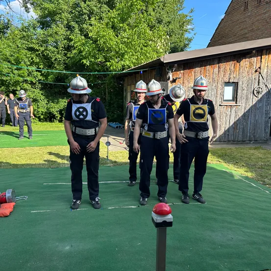Sechs Feuerwehrleute, mit Helmen, stehen auf einem grünen Feld mit einem roten Knopf. Sie tragen Schutzwesten mit einem X-Symbol. Bäume und ein Gebäude im Hintergrund.
