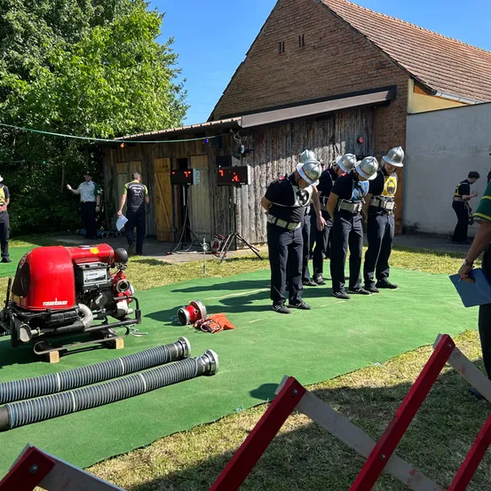 Eine Gruppe Feuerwehrleute in Helmen und Uniformen steht in einer Reihe auf grünem Rasen. Eine rote Maschine steht auf dem Boden mit langen Rohren. Ein Gebäude mit braunem Dach ist im Hintergrund.