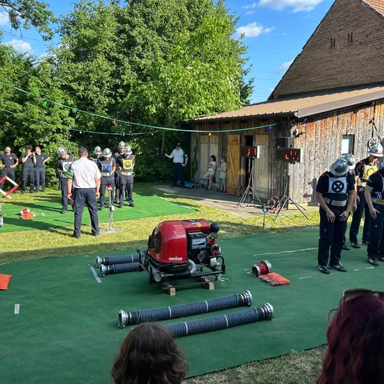 Eine Gruppe von Feuerwehrleuten trainiert auf einem grünen Feld mit einem Feuerwehrauto und Schläuchen vor einem Gebäude. Sie tragen Uniformen und Helme.