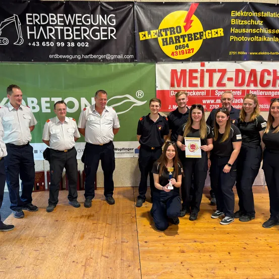 Eine Gruppe von Menschen in Uniform steht auf einem Holzboden. Eine Frau hält eine Trophäe, und zwei Banner stehen hinter ihnen. Eines sagt 'Erdbewegung Hartberger', und das andere sagt 'Meitz-Dach'.