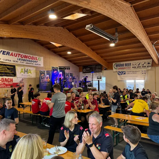 Eine Gruppe von Menschen versammelt sich in einem Veranstaltungsort mit Holztischen und -stühlen. Sie essen und einige klatschen. Der Veranstaltungsort hat eine Bühne mit einer Band und einer großen Leinwand. Banner hängen an den Wänden und Lampen sind an der Decke.