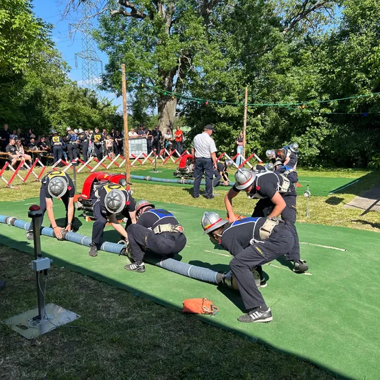 Eine Gruppe von Feuerwehrleuten trainiert auf einem grünen Feld. Sie tragen Helme und Uniformen. Es gibt eine Menschenmenge, die sie beobachtet.