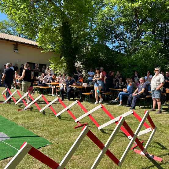 Eine große Menschenmenge versammelt sich in einem Außenbereich, einige sitzen auf Bänken, andere stehen. Im Vordergrund sind rote und weiße Barrieren aufgestellt, mit Bäumen und einem Gebäude im Hintergrund.