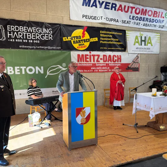 Ein Podium mit Schild und Mikrofon steht auf einer Bühne. Ein Mann spricht am Podium, begleitet von einem Priester und einer Frau am Klavier. Banner hängen im Hintergrund.