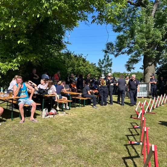 Menschen sitzen an Tischen in einem Park, einige tragen Hüte und Sonnenbrillen. Polizisten stehen im Hintergrund, und Barrieren sind in der Nähe der Bäume aufgestellt.