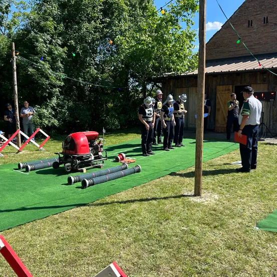 Eine Gruppe von Menschen in Helmen steht auf einem grünen Teppich in einem Feld, mit einer roten Maschine und Rohren in der Nähe. Ein Gebäude und Bäume sind im Hintergrund.