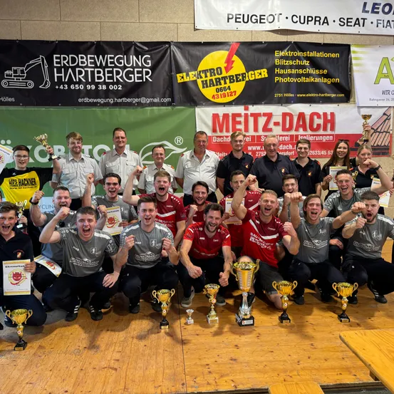 Eine Gruppe von Menschen posiert mit Trophäen, trägt verschiedene Shirts. Der Schauplatz scheint ein Saal mit Holzboden und Bannern zu sein.