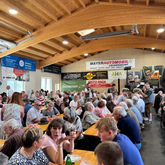 Eine Menge älterer Menschen sitzen auf Tischen in einem Innensaal, möglicherweise bei einem Festival. Einige klatschen, andere unterhalten sich. Banner mit Text hängen an den Wänden.