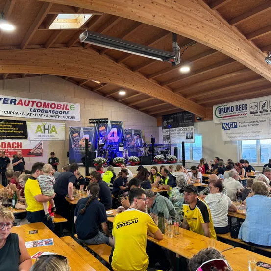 Eine große Gruppe von Menschen in gelben und blauen T-Shirts sitzt an Tischen in einem Saal. Banner an der Wand werben für eine Automarke und andere Sponsoren. Eine Bühne mit Blumen und einem Scheinwerfer ist hinten.
