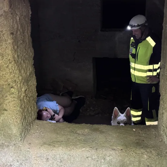 Eine Frau steckt in einem Loch fest, trägt ein blaues T-Shirt, und ein Hund ist in der Nähe. Ein Mann in Schutzkleidung steht mit einer Taschenlampe daneben.