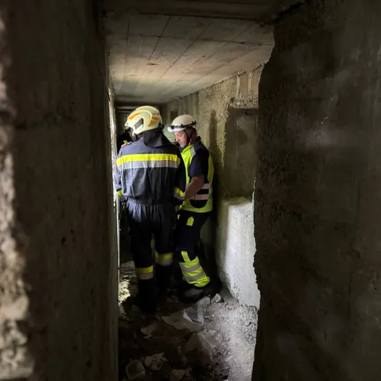 Zwei Arbeiter in Schutzkleidung gehen durch einen engen Betontunnel. Der Tunnel hat raue, unfertige Wände und einen schmutzig-erdigen Boden.