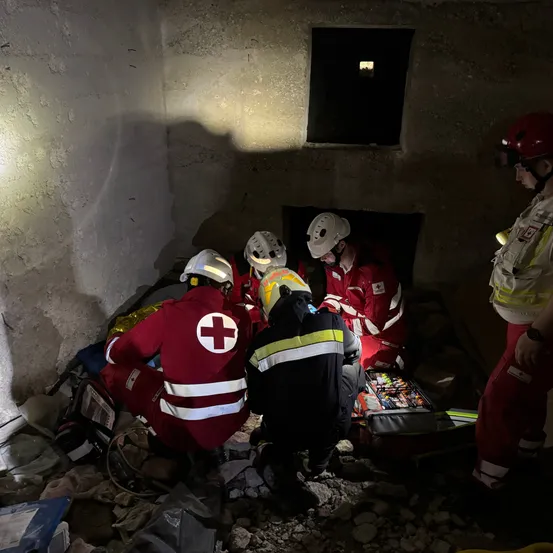 Rettungskräfte in roten und weißen Uniformen arbeiten in einem beschädigten Raum mit Geräten und Werkzeugen. Eine Person steht in der Nähe und beobachtet.