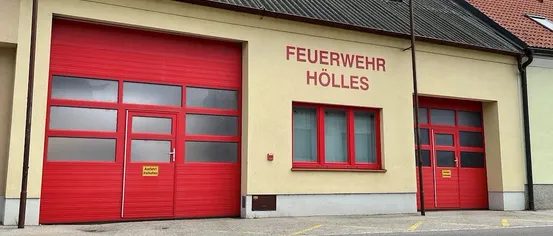 Die Vorderseite einer Feuerwache mit roten Türen und Fenstern. Ein Schild an der Wand lautet 'FEUERWEHR HÖLLES'. Rechts befindet sich ein Pfosten mit einer Flagge.