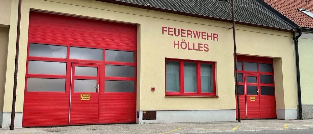 Die Vorderseite einer Feuerwache mit roten Türen und Fenstern. Ein Schild an der Wand lautet 'FEUERWEHR HÖLLES'. Rechts befindet sich ein Pfosten mit einer Flagge.