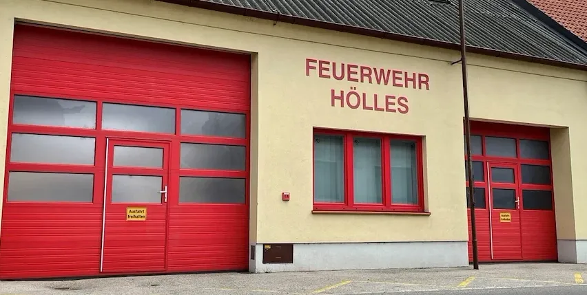 Die Vorderseite einer Feuerwache mit roten Türen und Fenstern. Ein Schild an der Wand lautet 'FEUERWEHR HÖLLES'. Rechts befindet sich ein Pfosten mit einer Flagge.