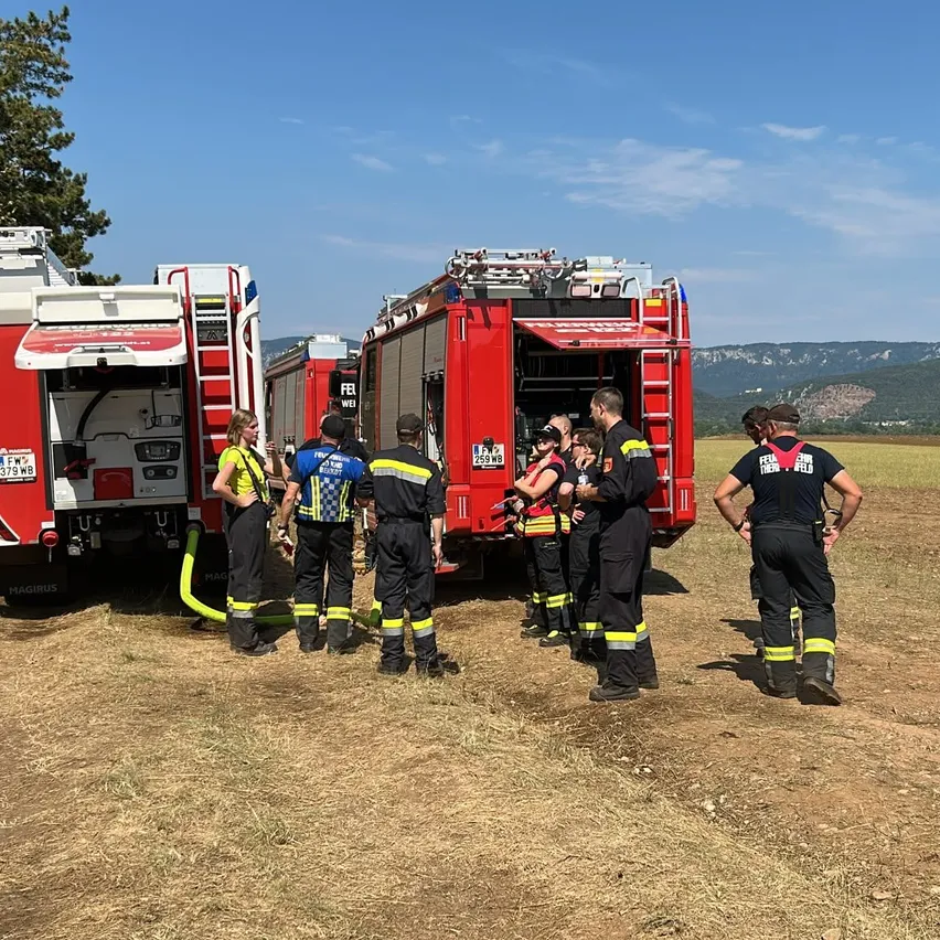Eine Gruppe von Feuerwehrleuten steht um einen Feuerwehrwagen herum, einer von ihnen hält eine Feuerwehrschläuche. Sie tragen Uniformen und Helme. Hinter ihnen stehen zwei weitere Feuerwehrwagen. Der Himmel ist klar mit einigen Wolken. In der Ferne gibt es Berge.