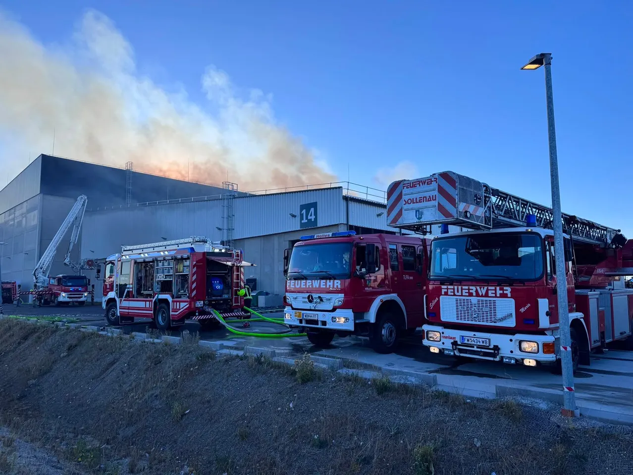 Drei rote Feuerwehrfahrzeuge sind vor einem Gebäude geparkt, in dessen Hintergrund Rauch aufsteigt. Ein Fahrzeug hat eine Leiter auf dem Dach.