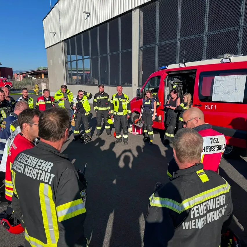 Eine Gruppe von Feuerwehrleuten in gelben und schwarzen Uniformen steht um einen roten Feuerwehrwagen mit der Aufschrift HLF1 herum. Sie stehen vor einem Gebäude mit Glasfenstern.