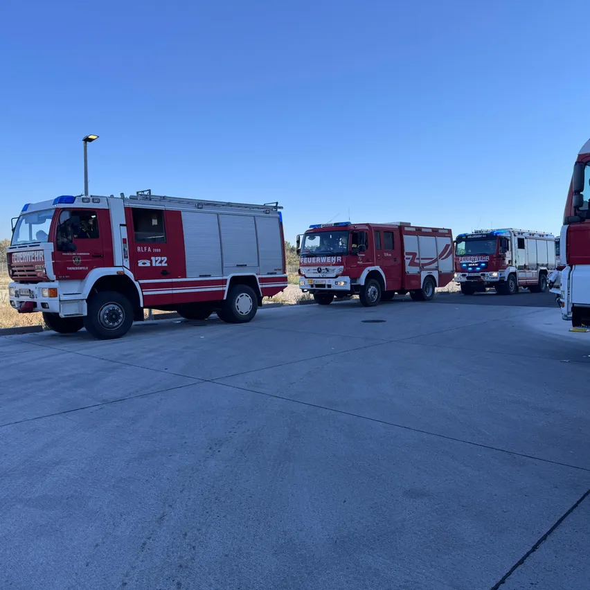Vier Feuerwehrfahrzeuge sind in einer Reihe auf einer Betonfläche geparkt, mit einem klaren blauen Himmel im Hintergrund.