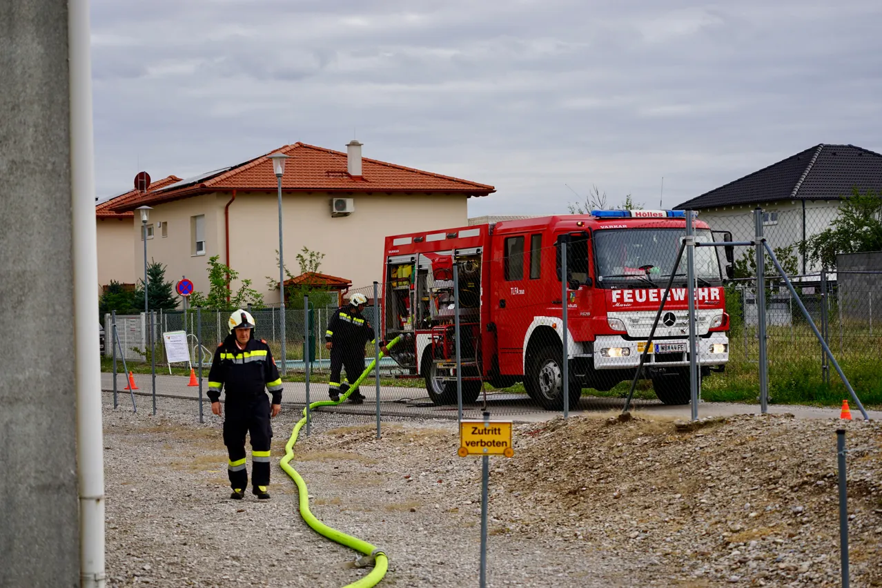 Zwei Feuerwehrleute in voller Ausrüstung bedienen einen roten Feuerwehrwagen mit einer angeschlossenen Schlauchleitung. Der Wagen steht vor einem Haus mit rotem Dach.