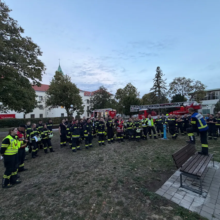 Eine Gruppe von Feuerwehrleuten versammelt sich in einem Park, mit einem Feuerwehrauto im Hintergrund. Sie sind in ihren Uniformen gekleidet und scheinen einer Einweisung zuzuhören.