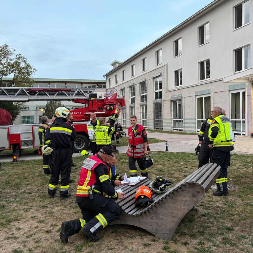 Eine Gruppe von Feuerwehrleuten in Uniform versammelt sich um eine Bank in einem Grasbereich, mit einem Feuerwehrwagen im Hintergrund.