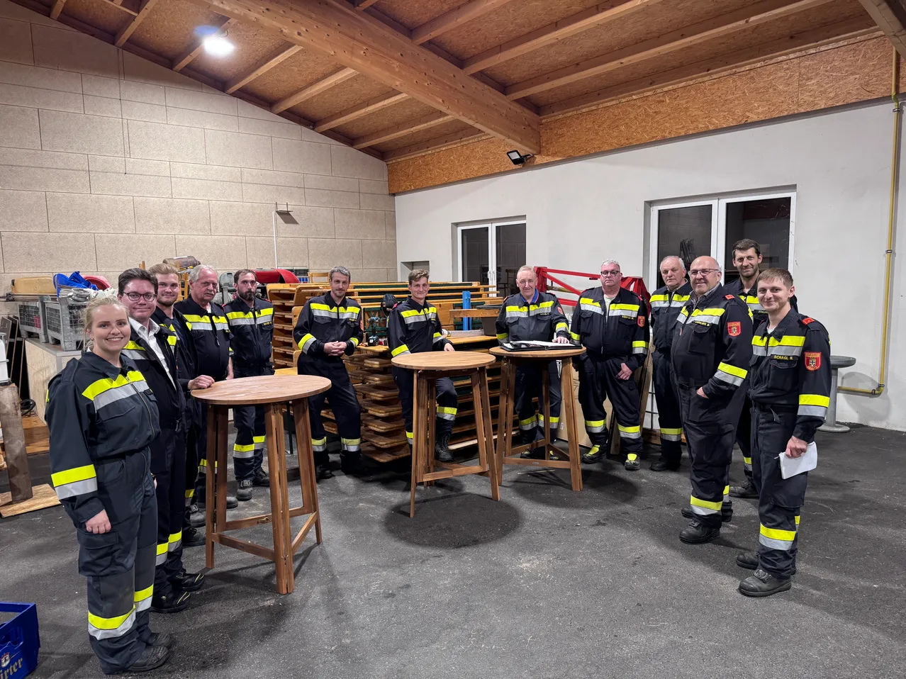 Eine Gruppe von Feuerwehrleuten in Uniform steht um Holztische in einem Lagerhaus. Sie sind für eine Besprechung oder Veranstaltung versammelt.