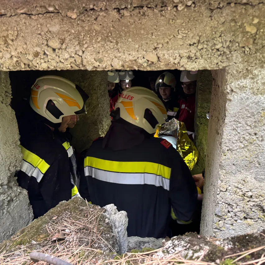 Feuerwehrleute in Schutzausrüstung arbeiten in einem engen Raum, möglicherweise einem Tunnel, um jemanden zu retten. Sie sind mit Helmen ausgestattet und einer hält eine silberne Folie.