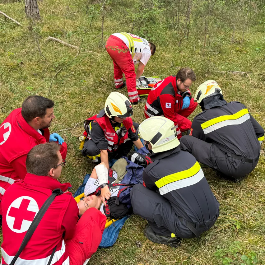 Mehrere Rettungssanitäter in roten und gelben Uniformen kümmern sich um einen Patienten auf einer Trage in einem Grasfeld. Sie sind mit Helmen und Schutzausrüstung ausgestattet.
